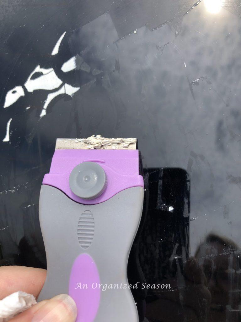 A hand holding a razor that is scrapping a window decal off of a car window. A tip for how to organize and clean your car.