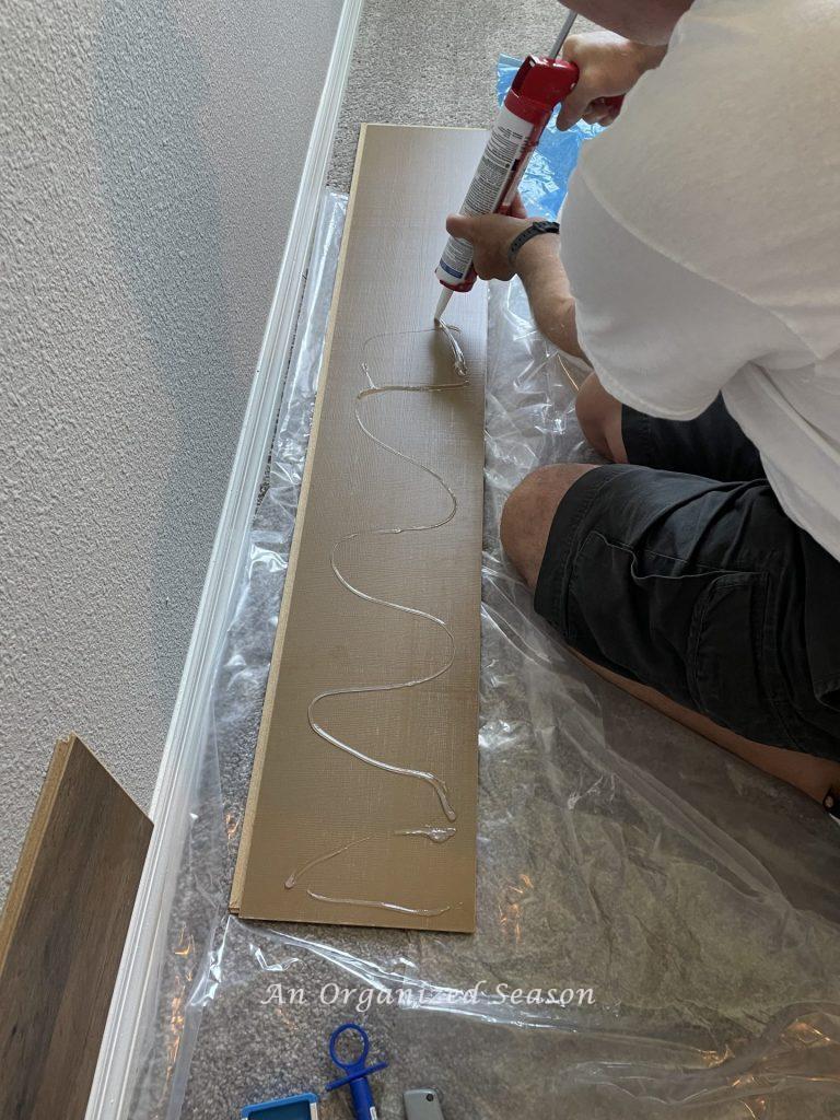 A man applying silicone to the back of a piece of wood flooring. Step three for how to install flooring on your wall.