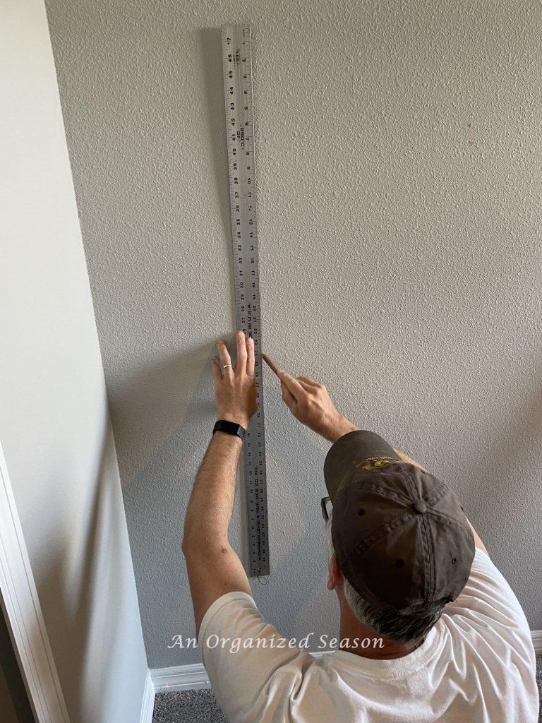 A man using a long metal ruler to draw vertical lines on a wall. Step three of advice to install flooring on a wall.