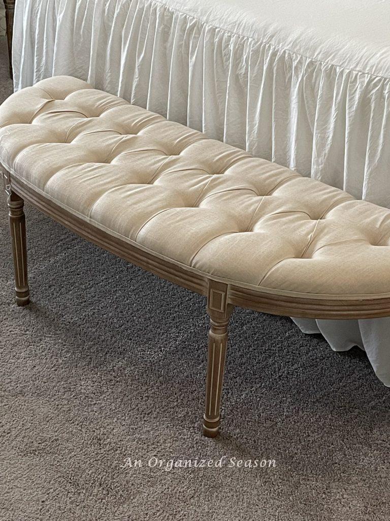 A cream tufted bench at the end of a bed. 