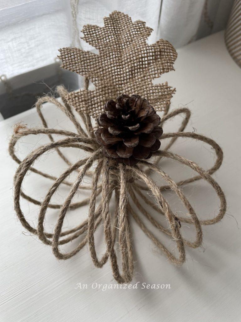 A twine pumpkin embellished with a pinecone stem and burlap leaf. One example of how to make two types of twine pumpkins.