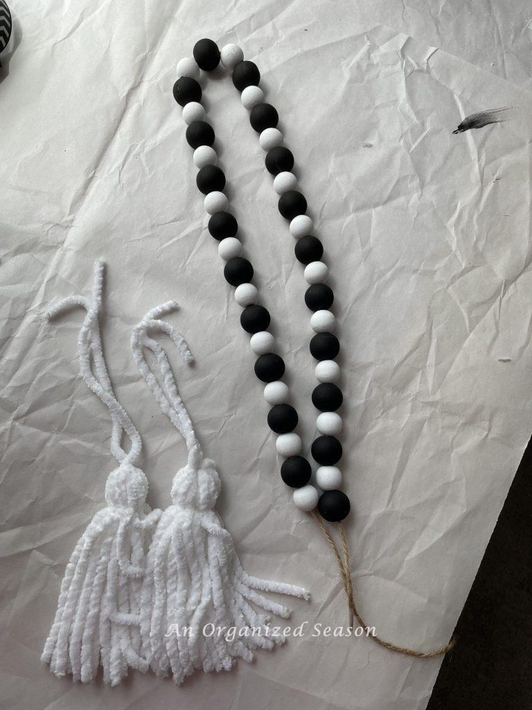 A black and white wood bead garland with two white tassels next to it. Showing how to make a Halloween wood bead garland.