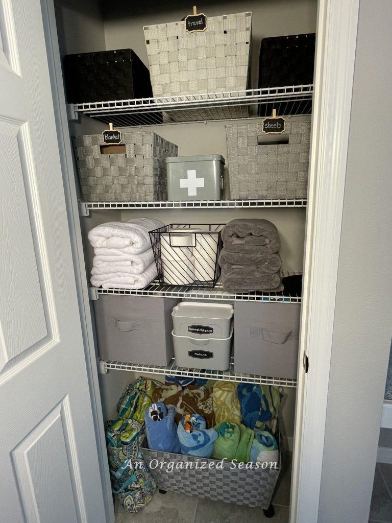 A master bathroom closet with gray, white, and black containers storing various items. Bath towels are folded on one shelf and pool towels are rolled and stored in a basket on the floor. Examples of master bathroom organization. 
