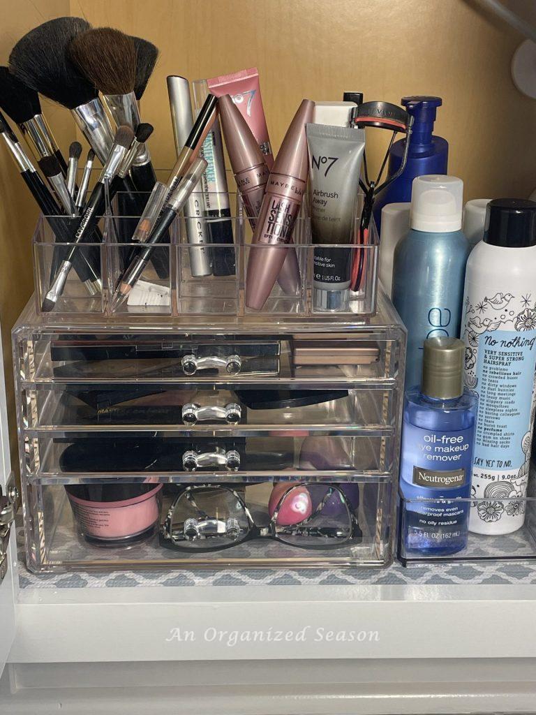 Clear plastic storage container holding make-up and brushes inside a bathroom cabinet.  An example of master bathroom organization. 