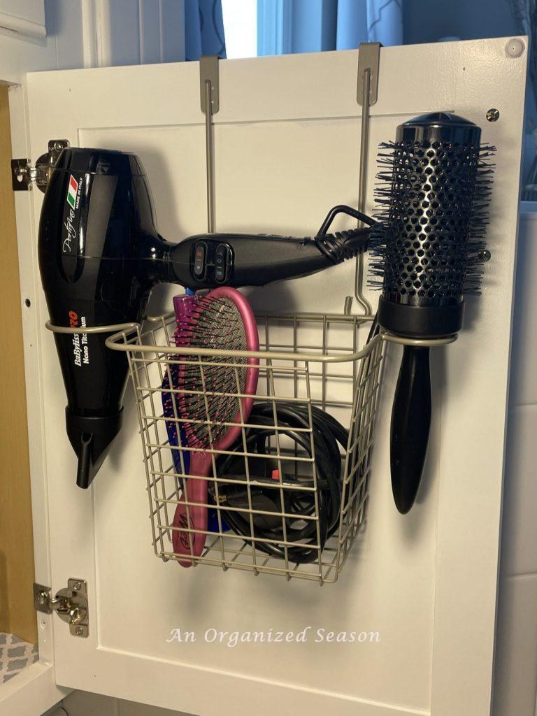A basket hanging on the back of a bathroom cabinet door filled with hair products. An example of master bathroom organization.