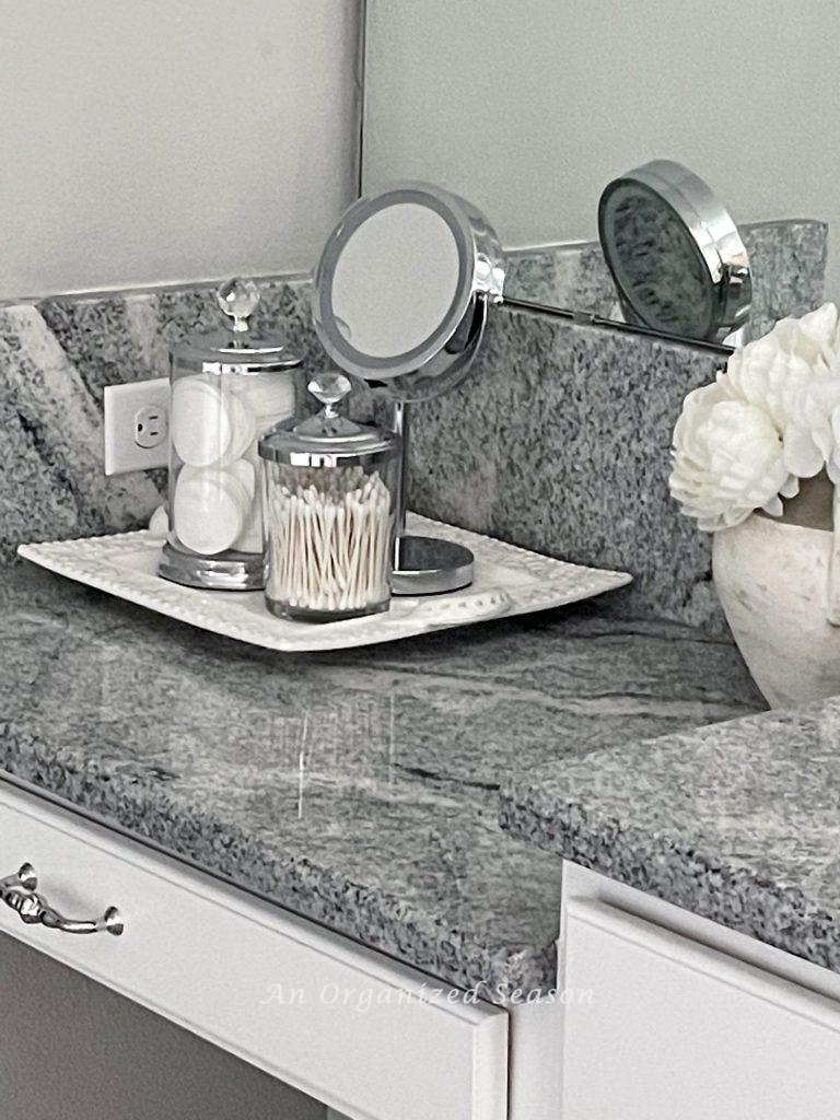 Two clear glass lidded containers holding cotton pads and swabs and a make-up mirror sitting on a white tray on a bathroom counter. An example of master bathroom organization ideas! 