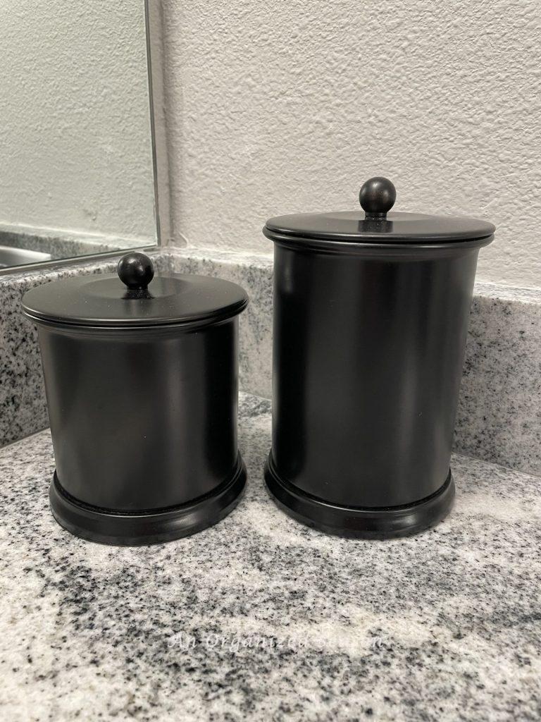 Two black canisters in different heights storing items on a bathroom counter.  An example of master bathroom organization ideas. 