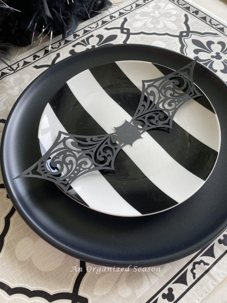 A black and white striped salad plate sitting on top a black dinner plate with a cut out shape of a bat on top. An example of inexpensive Halloween decor for your kitchen.