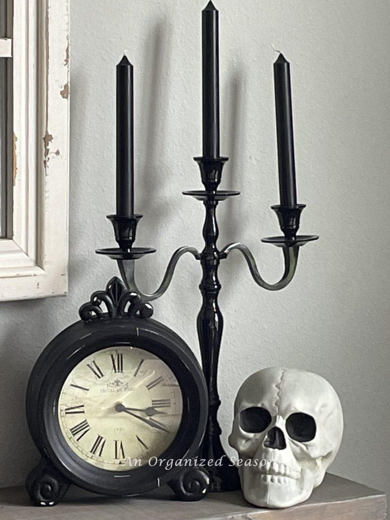A black candle holders with three black candles next to a black clock and a skull.  An example of inexpensive Halloween decor for your kitchen.