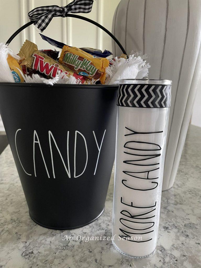 A Rae Dunn inspired candle that says "more candy" sitting beside a black bucket of Halloween candy. An example of a Rae Dunn inspired candle you can learn how to make! 