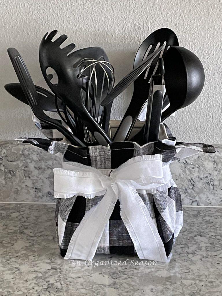 A kitchen utensil holder that has been wrapped with a black and white buffalo check napkin and tied with a white bow., holding black kitchen utensils. An example of inexpensive Halloween decor for your kitchen.