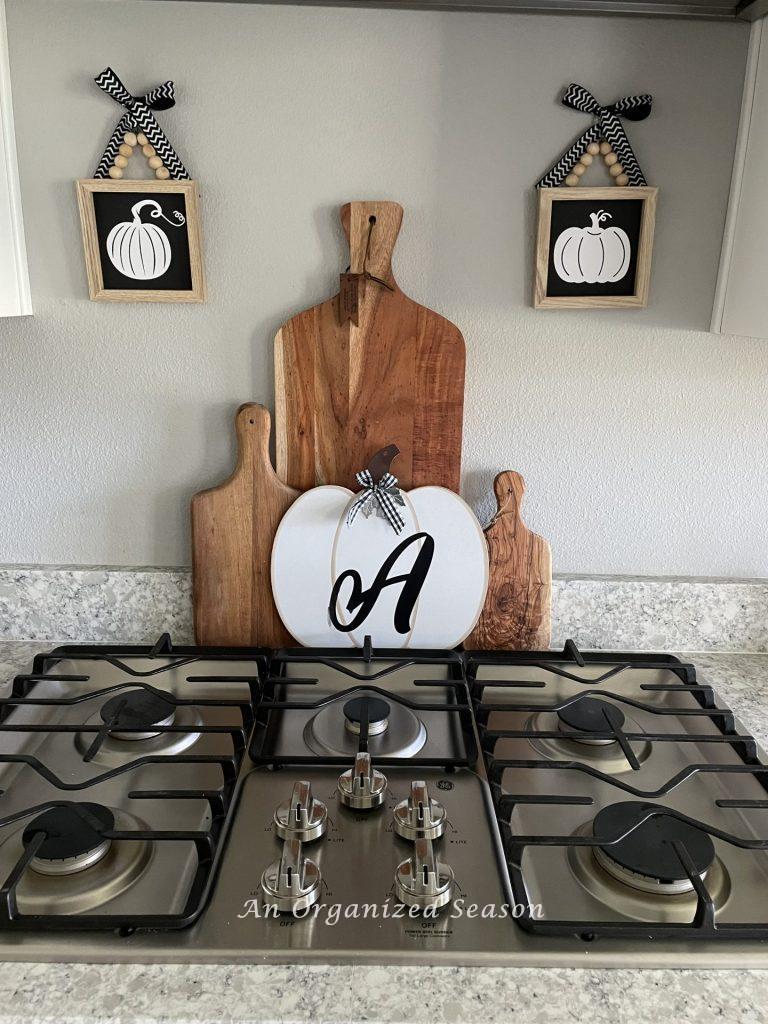 Three wooden cutting boards and a white wood pumpkin with the letter "A" printed on it are stacked on a kitchen counter behind the stove. Black and wwhite pumpkin pictures hang on each side, an example of inexpensive Halloween decor for your kitchen.