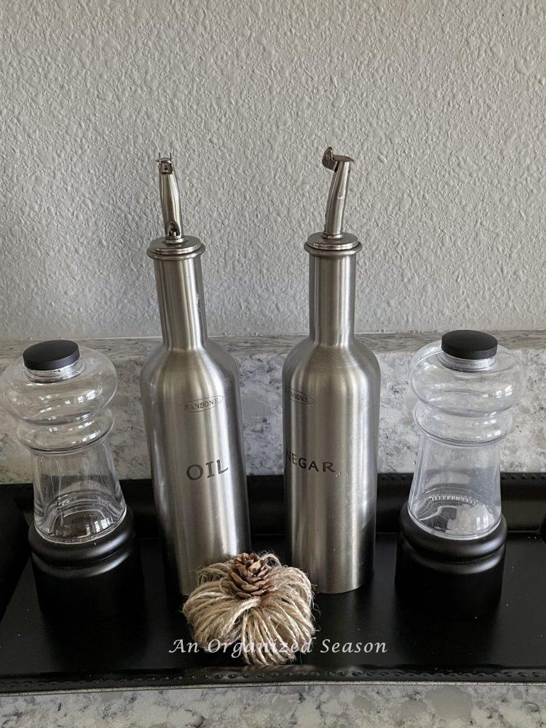 Silver oil and vinegar dispensers on a black tray with salt and pepper grinders and a small twine pumpkin. An example of inexpensive Halloween decor for your kitchen. 