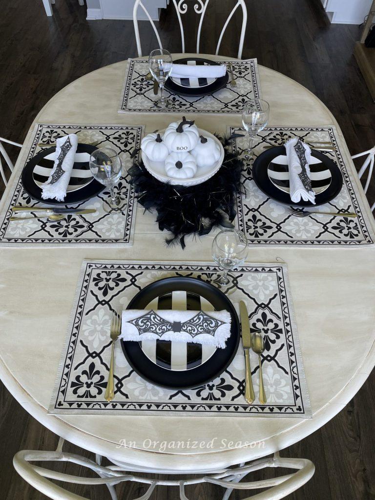 A white kitchen table set with four black and white place settings and a white pumpkin centerpiece. An example of inexpensive Halloweeen decor for your kitchen.