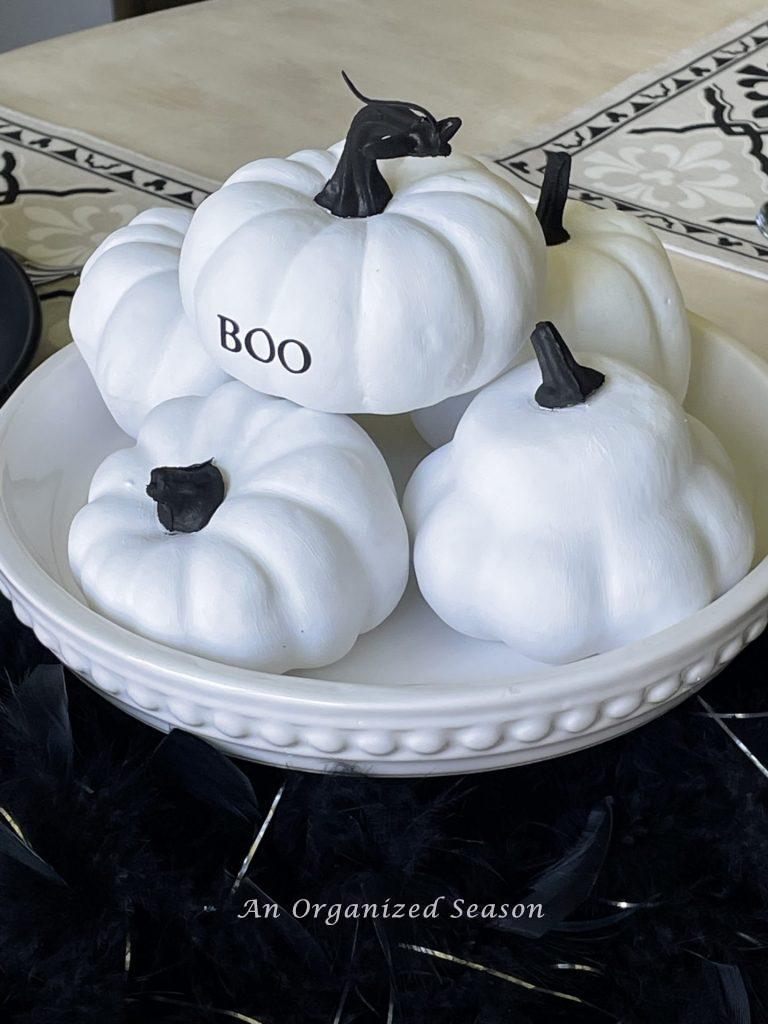 A white cake stand stacked with white pumpkins that have black stems. The top pumpkin has the word "BOO" written on it. Black feathers surround the stand of the cake plate. An example of inexpensive Halloween decor for your kitchen.