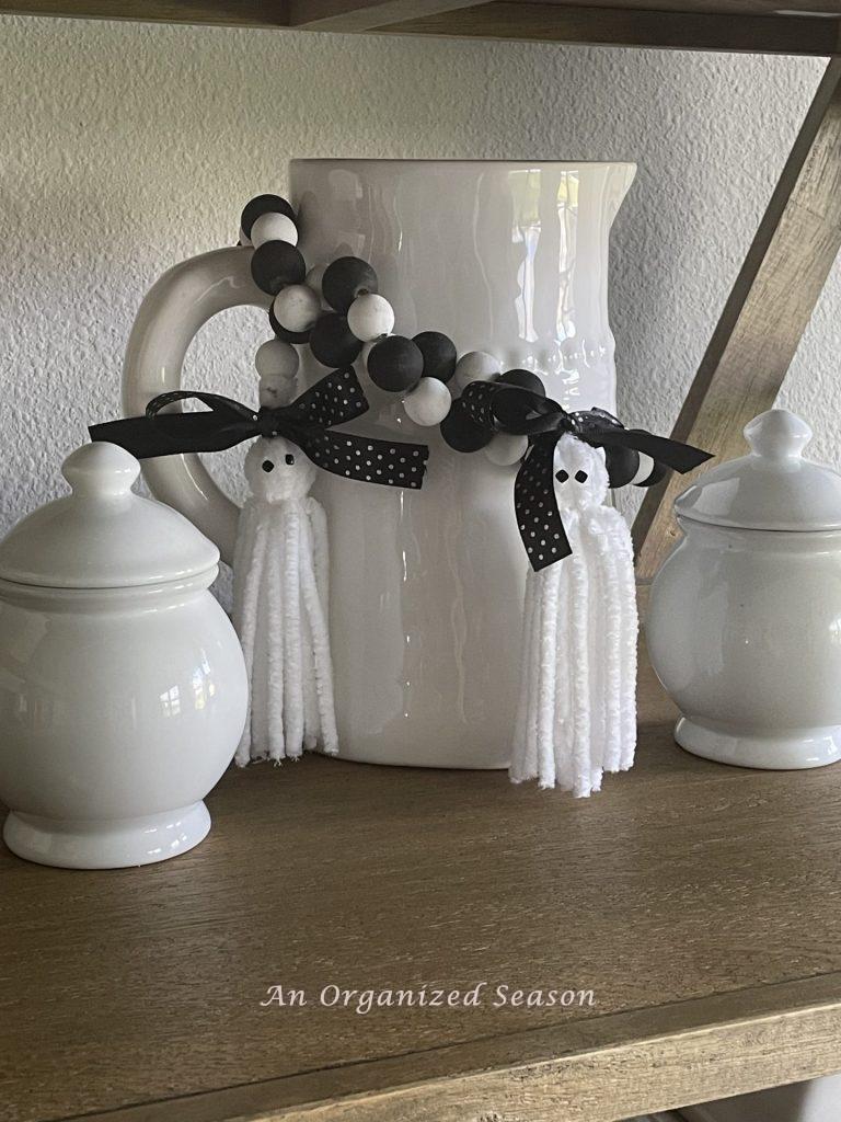 A black and white wood bead garland with ghost tassels, wrapped around a white pitcher with two white covered containers. An example of inexpensive Halloween decor for your kitchen.