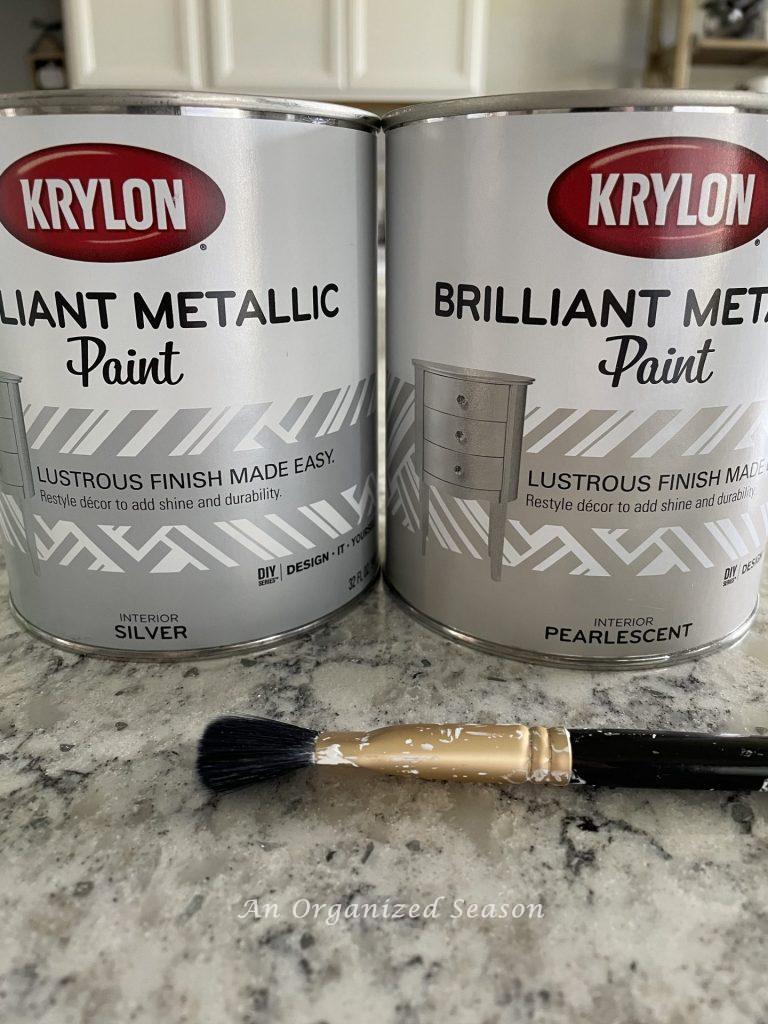 Two cans of Krylon Brilliant Metallic Paint in Silver and Pearlescent and a paint brush. Items used in how to apply pearlescent paint on pumpkins. 