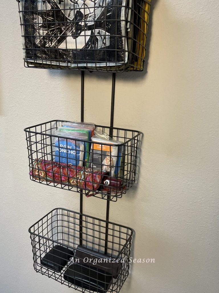 A three-tiered wire basket hanging on a wall, holding reusable grocery bags, ponchos, umbrella, sunglasses case and a wallet. An example of how to organize every entryway to your home.