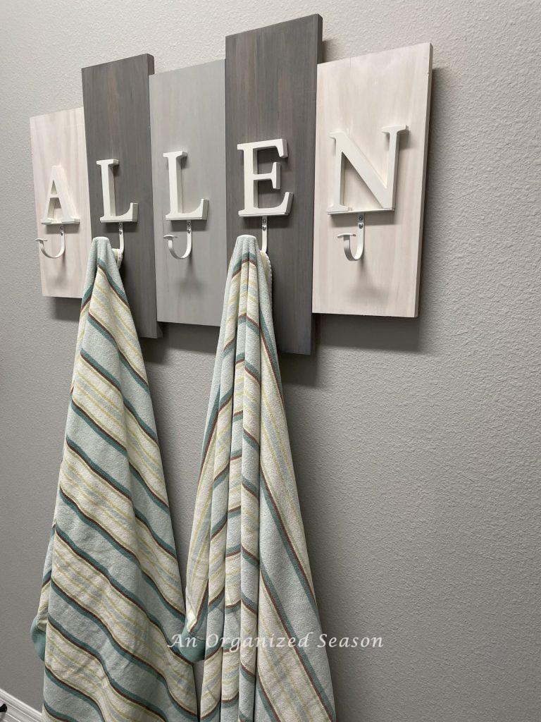 A gray and white wooden coat rack with hooks that spell "ALLEN". Two beach towels are hanging from the hooks. An example of how to organize the entryway to your home. 