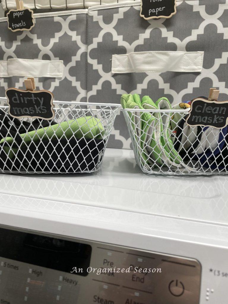 Two small white wire baskets sitting on a washing machine. Each has a chalkboard tag that says "dirty masks" and "clean masks". An example of how to organize the entryway to your home.