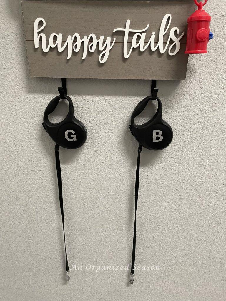 A gray wood sign with white letters spelling "happy tails". Sign has two hooks below it holding two black dog leashes. An example of how to organize the entry way to your home.