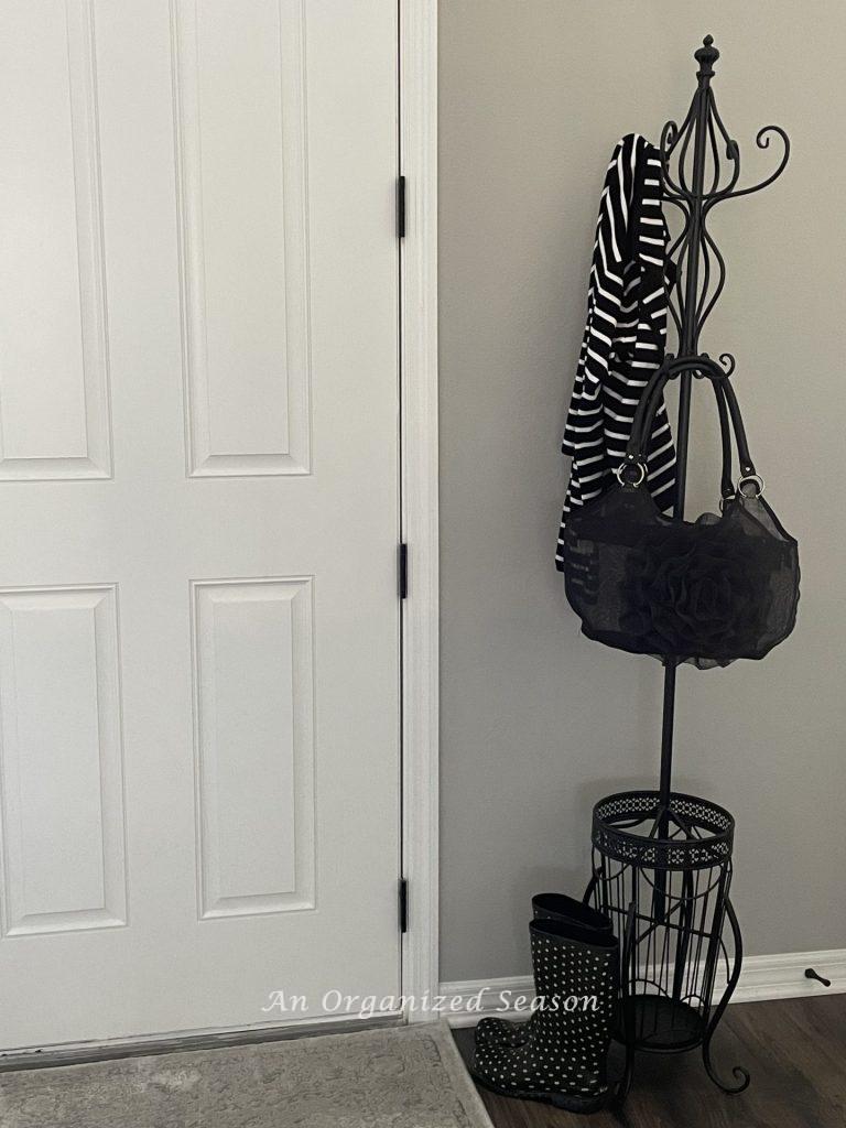 A black metal coatrack with built-in umbrella stand. Black purse and stripped jacket are hanging on it and black and white polk-a-dot rain boots are sitting beside it. An example of how to organize the entryway to your home.