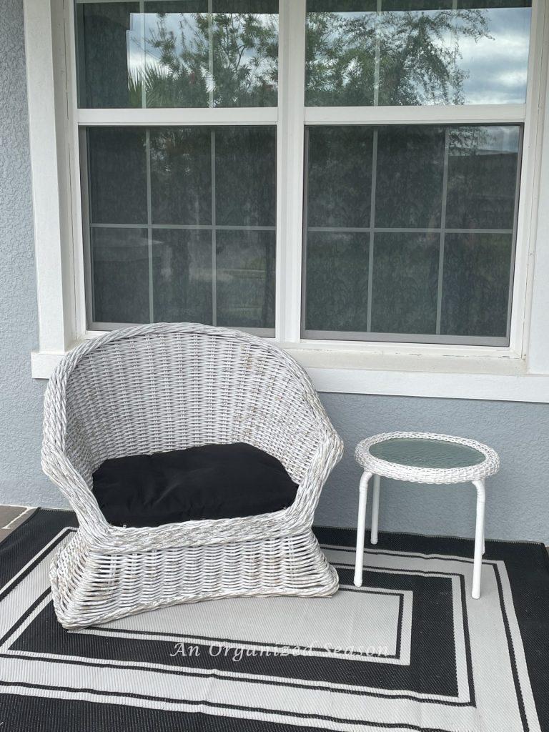 A front porch with black and white rug, white wicker chair, and table. An example of how to decorate your front porch for Fall.