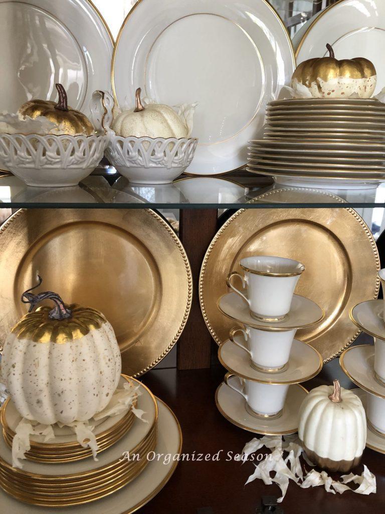 An organized china cabinet with ivory and gold dishes and white and gold Fall pumpkins. An example of how to set Fall home organization goals.
