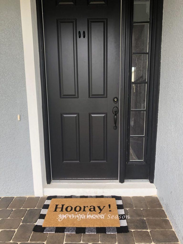 A front porch with black door and buffalo check rug layered with a tan doormat, showing how to decorate a front porch for Fall. 