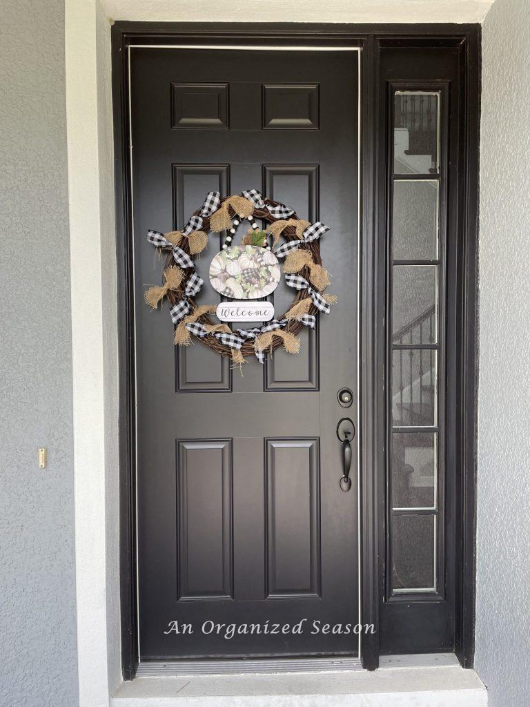 A fall wreath decorated with buffalo check and burlap bows, hanging on a black door. Showing how to decorate a porch for Fall. 