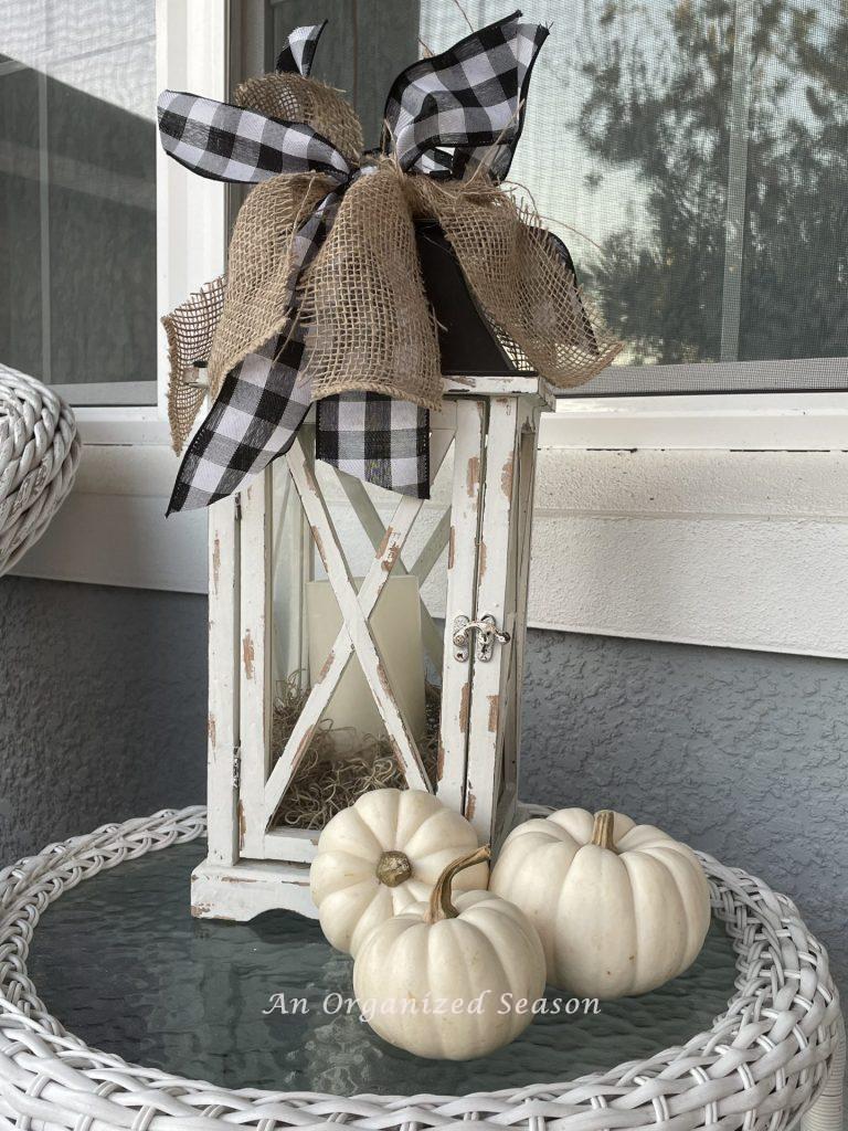 A white lantern with buffalo check and burlap ribbon sitting on a white wicker table next to three white pumpkins. Showing how to decorate a porch for Fall. 