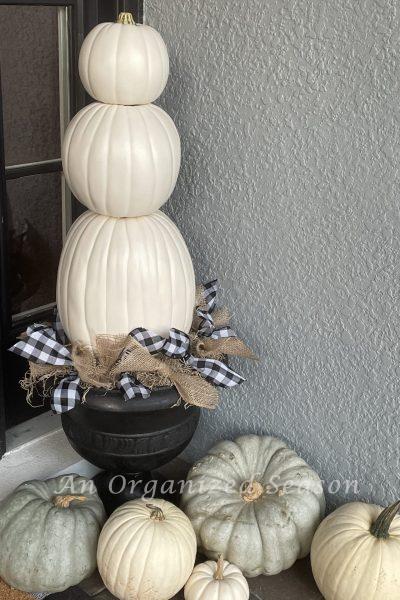 A three-tiered white pumpkin topiary in a black planter surrounded by white and green heirloom pumpkins