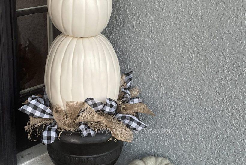 A three-tiered white pumpkin topiary in a black planter surrounded by white and green heirloom pumpkins