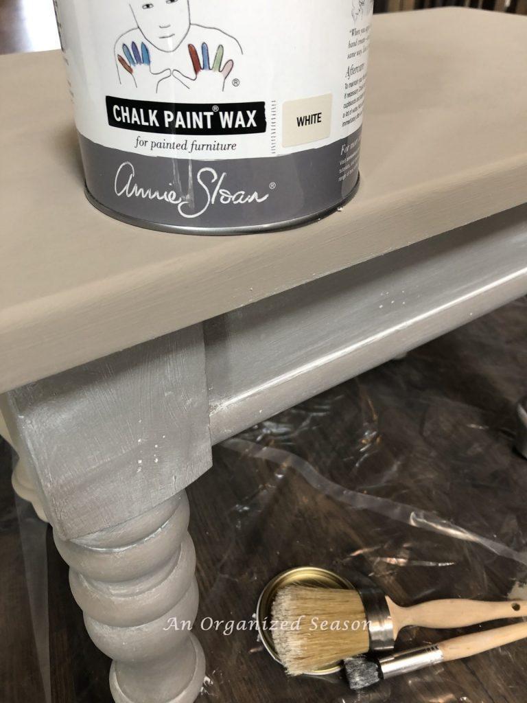 A wood table with a can of Annie Sloan Wax in white sitting on top of it. Top of table looks beige and half of table looks gray after the wax was applied.  Step three for my living room table makeover.