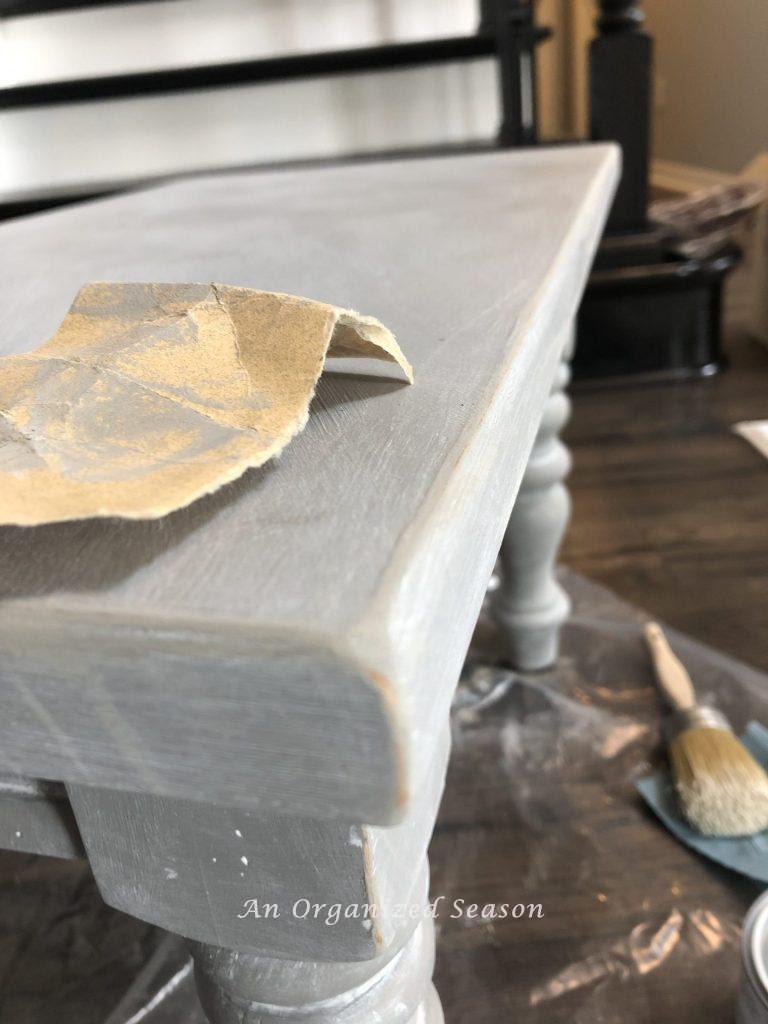 A table that has been chalk painted, waxed, and distressed with the piece of sandpaper sitting on top of it. Step four of my living room table makeover.