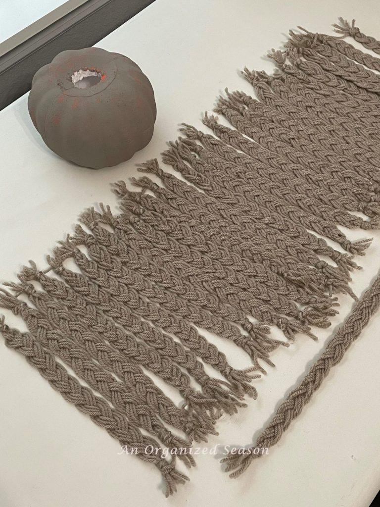 A gray faux pumpkin with holes in the top and bottom next to 32 braided strands of tan yarn, used to make an adorable yarn pumpkin.