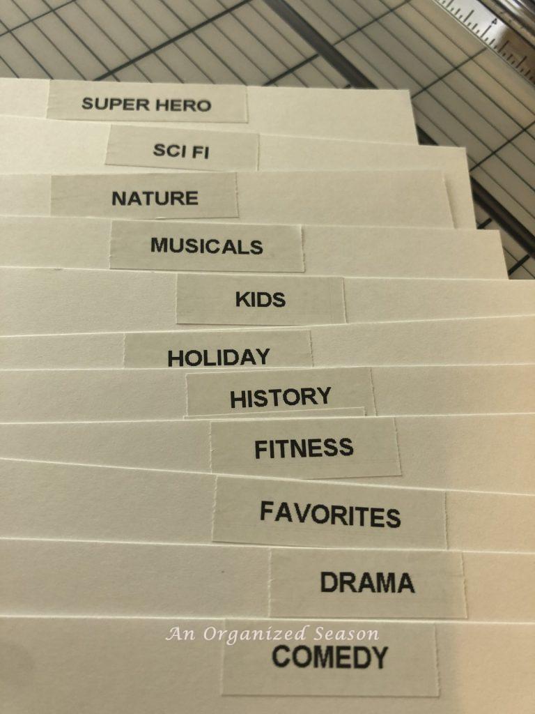 Pieces of white cardstock with labels attached to the top with different movie genres that will be used to organize the DVDs. 