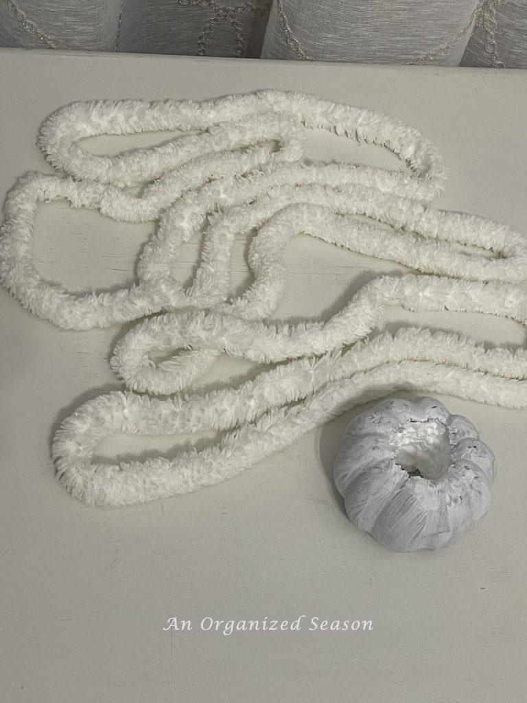 A six foot strand of chenille yarn next to a faux pumpkin with a hole in top and bottom. Items needed to make an adorable yarn pumpkin. 