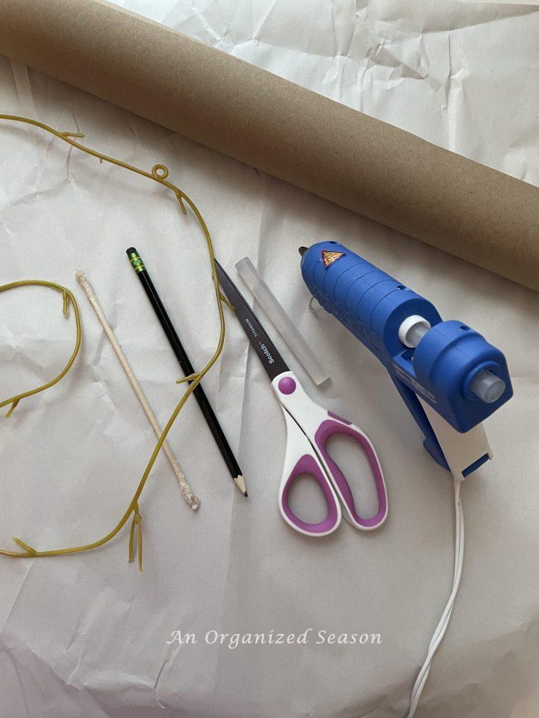 A roll of brown craft paper, a glue gun, glue stick, pencil, scissors, and orangewood stick, items needed to create a simple paper leaf garland.
