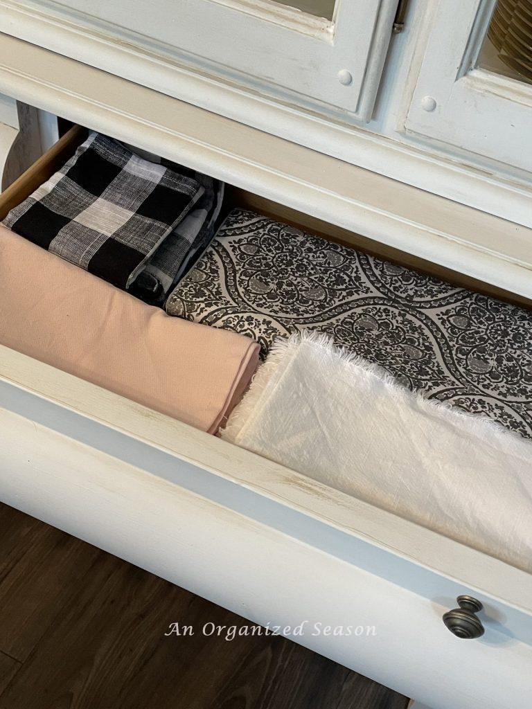 An open drawer in a china cabinet holding cloth napkins and a table runner. An example of how to organize your dining room for easy entertaining.