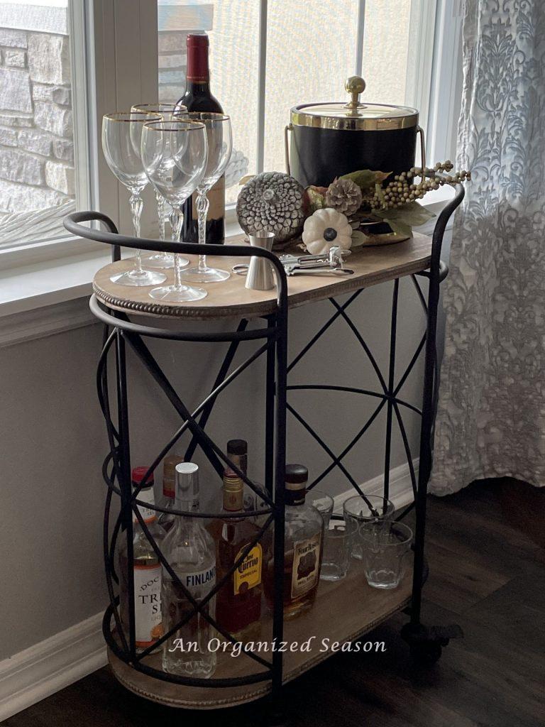 An organized bar cart set up in a dining room with wine glasses, ice bucket, wine bottle opener, several bottles of liquor, and highball glasses. An example of how to organize your dining room for easy entertaining. 