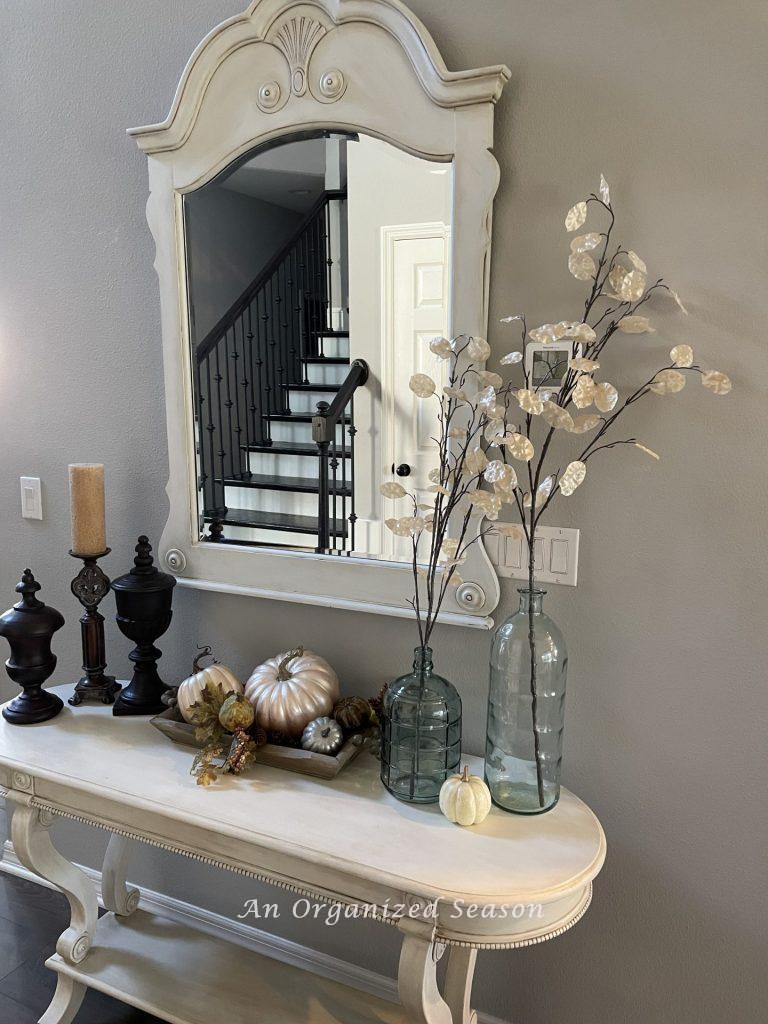 A table and mirror in an entryway, decorated for Fall with stems of neutral leaves and muted pumpkins. The first room of the best of Autumn home tour.