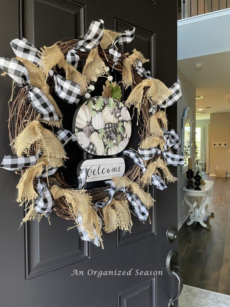 The front door of a home opened into the foyer. The door has a black, white, and green fall wreath hanging on it, welcoming people to the best of Autumn home tour. 