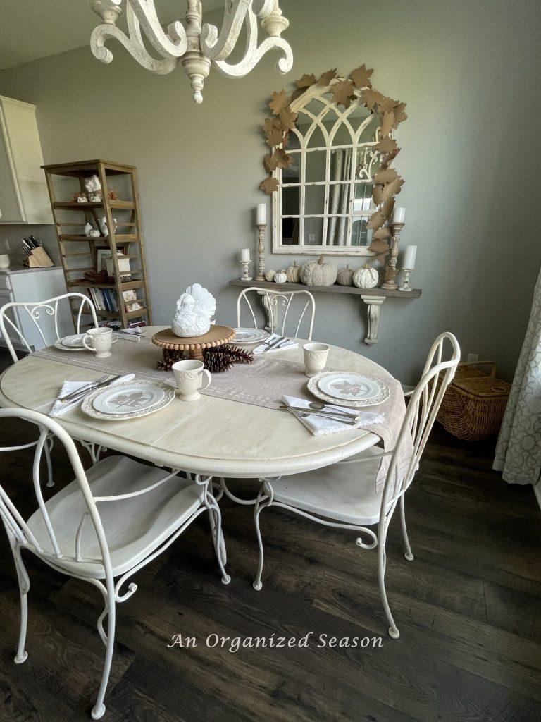An eat-in kitchen table decorated in neutral place settings with a white turkey centerpiece. Showing room five of the best of Autumn home tour.