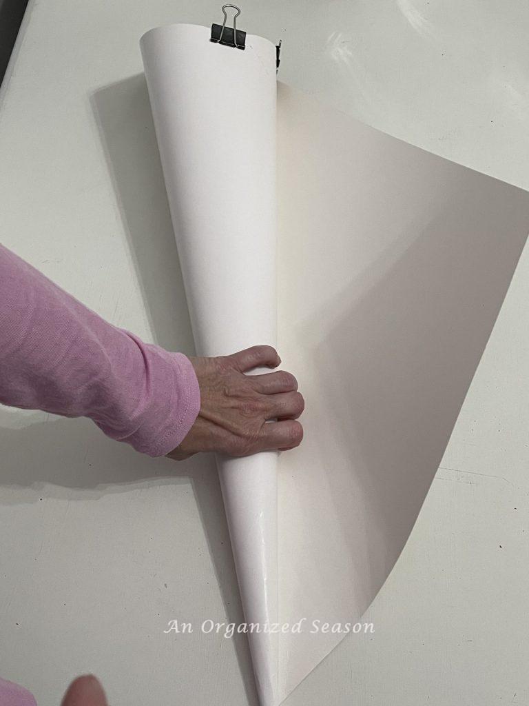 A womans hand rolling a white poster board in to a cone shape to make a DIY cone Christmas tree.