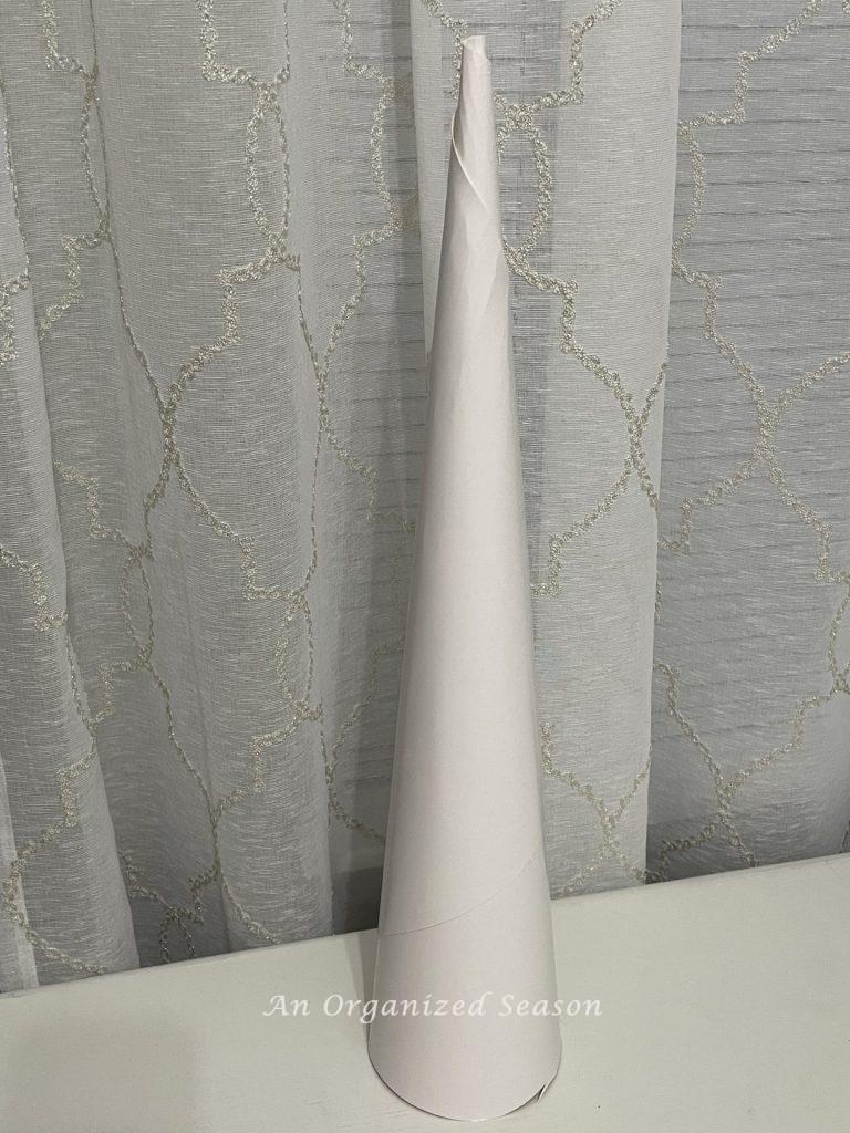A long white skinny poster board cone sitting on a table. An example of how to make a DIY cone Christmas tree.