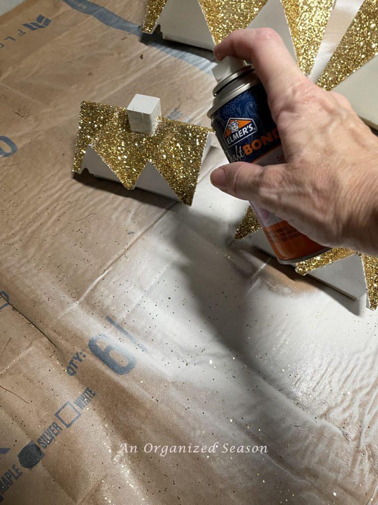 Step six to make a DIY Christmas village is to spray the glittered roof with spray adhesive. 