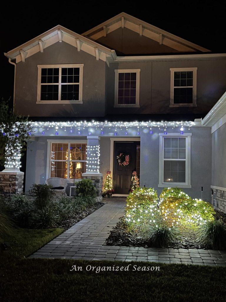 A house with white lights wrapped around columns and on the bushes. Part of the Tis the season nighttime home tour.