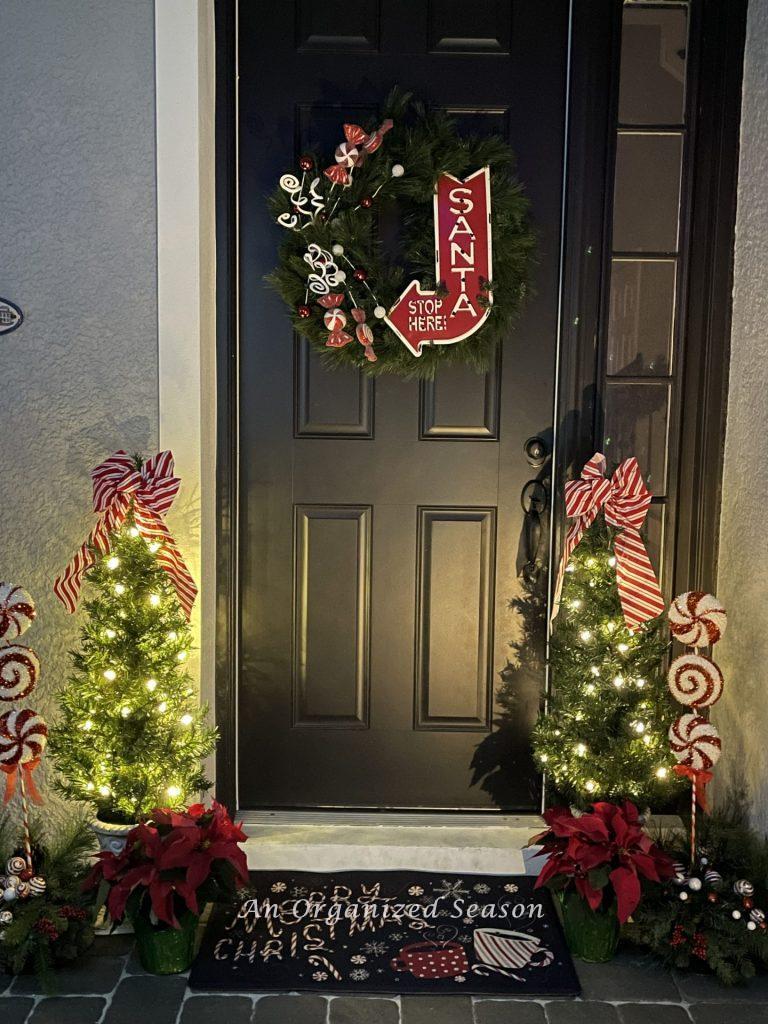 A front door surrounded by two small trees with white lights and candy striped bow during the Tis the Season nighttime home tour.