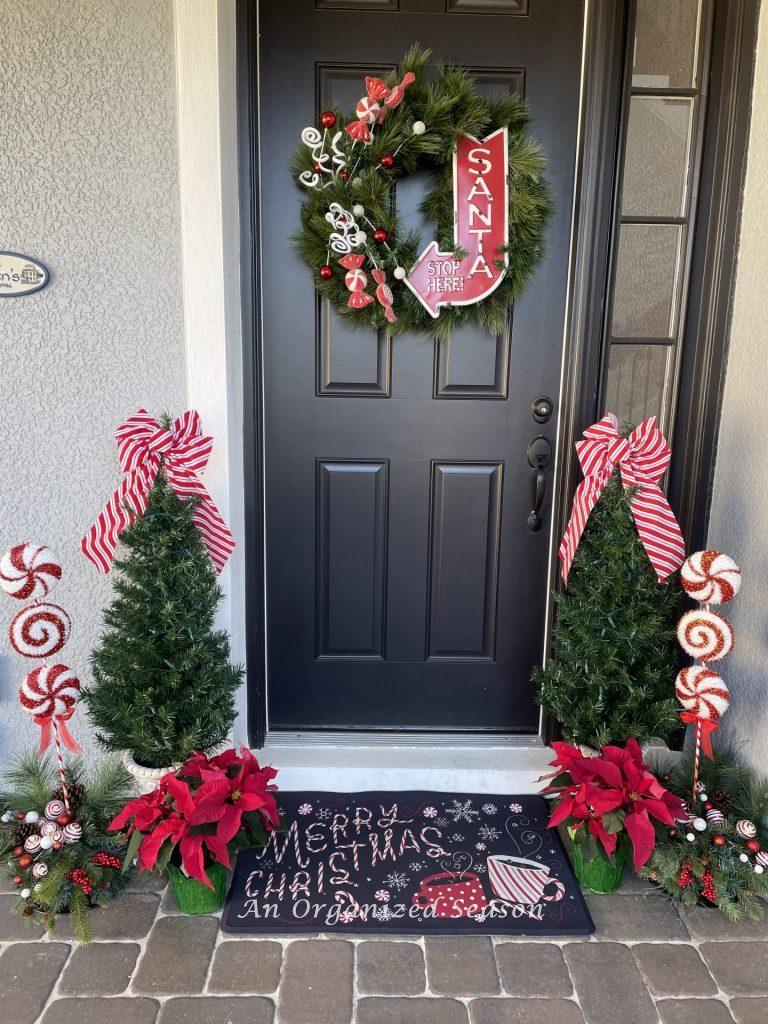 Holiday porch decor with a wreath, two Christmas trees, poinsettias, and rug. 